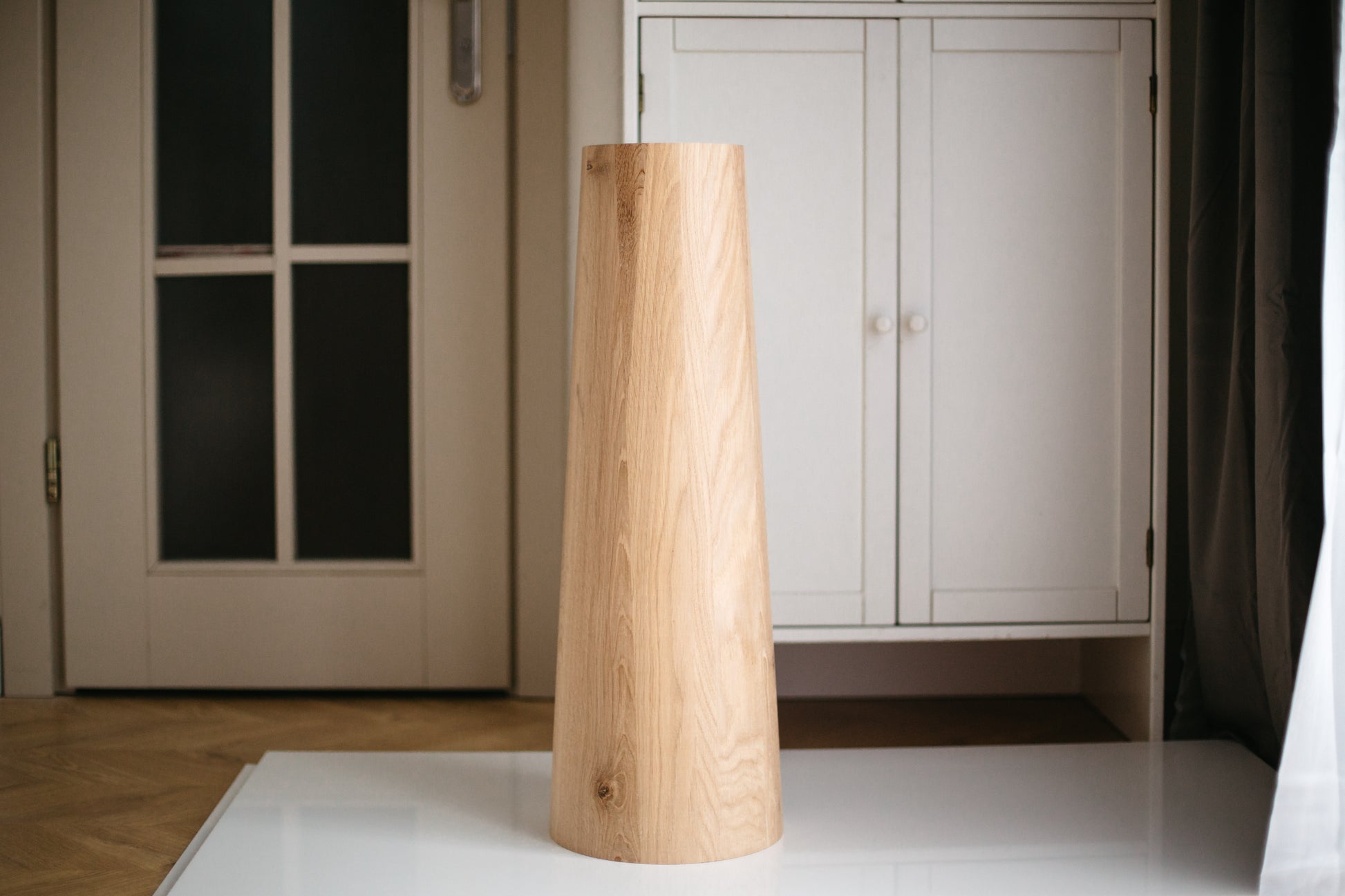 A single wooden furniture leg standing upright on a white surface, with a door and a wardrobe in the background.