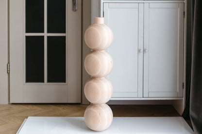 A tall, minimalist decorative vase made of four stacked, rounded wooden spheres in a light natural wood finish, standing on a white surface in an indoor room setting.