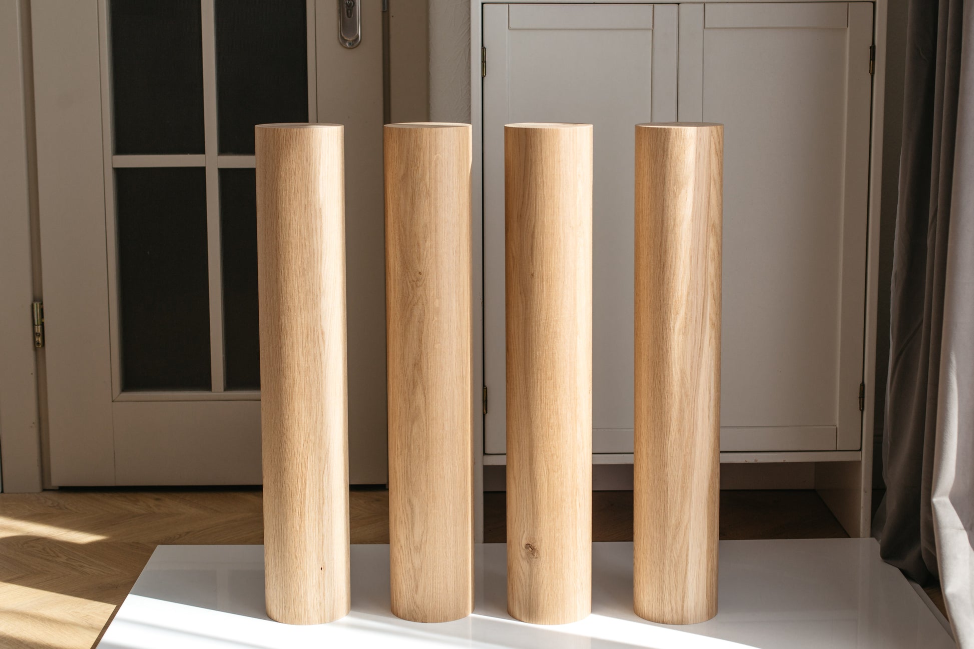 Four tall, cylindrical wooden poles or posts with a natural light oak finish, standing upright on a white surface in front of a white cabinet in a home interior.
