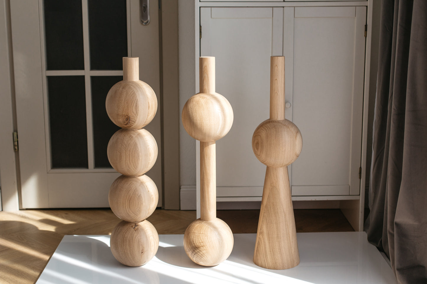 Three light natural wood decorative sculptures with rounded geometric shapes, displayed on a white table in a bright room with a white cabinet and dark curtain in the background.