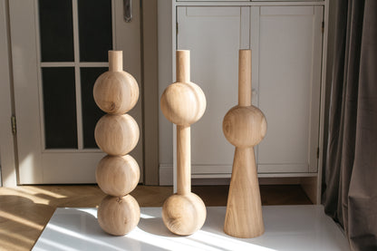 Three light natural wood decorative sculptures with rounded geometric shapes, displayed on a white table in a bright room with a white cabinet and dark curtain in the background.