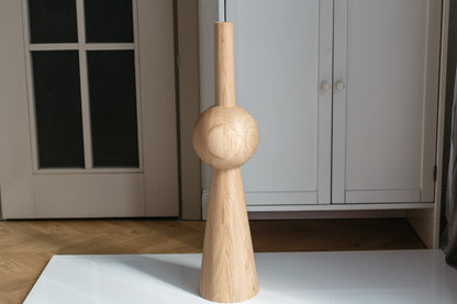 A light natural wood decorative object with a flared base, a rounded middle section, and a narrow top stem, standing on a white surface in an indoor room with cabinets and a door behind it.