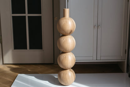A modern wooden decorative sculpture made of four stacked rounded spheres on a slim cylindrical top, standing on a white surface in a sunlit room with light gray cabinets in the background.
