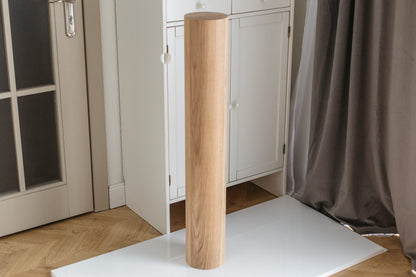 A tall, cylindrical wooden column or lamp base with a natural oak wood grain finish, standing upright on a white surface in a room setting with a white cabinet, gray curtain, and wooden floor.