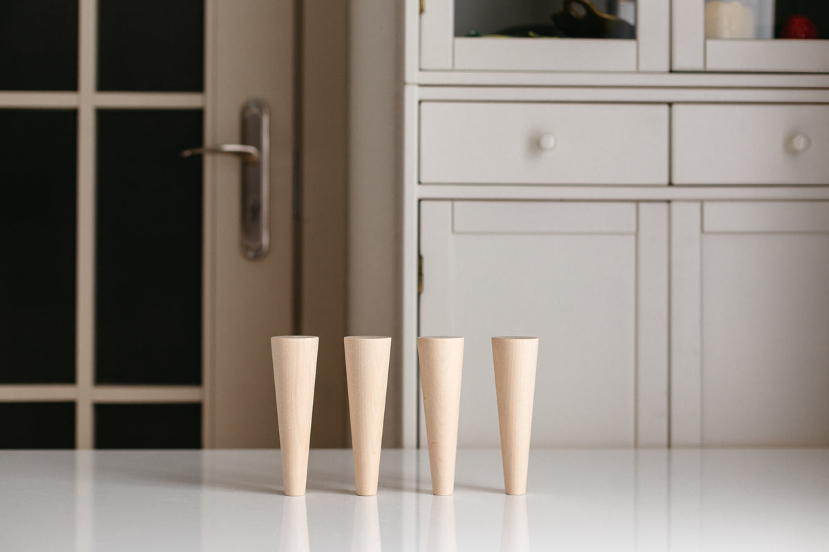 Four light natural wood tapered cups standing upright on a white tabletop, with a softly blurred indoor background featuring a cabinet and door.