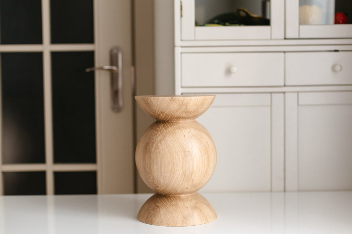 A small wooden pedestal stand with a rounded middle section and flared top and bottom bases, shown on a white tabletop with a blurred interior background featuring a cabinet and door.
