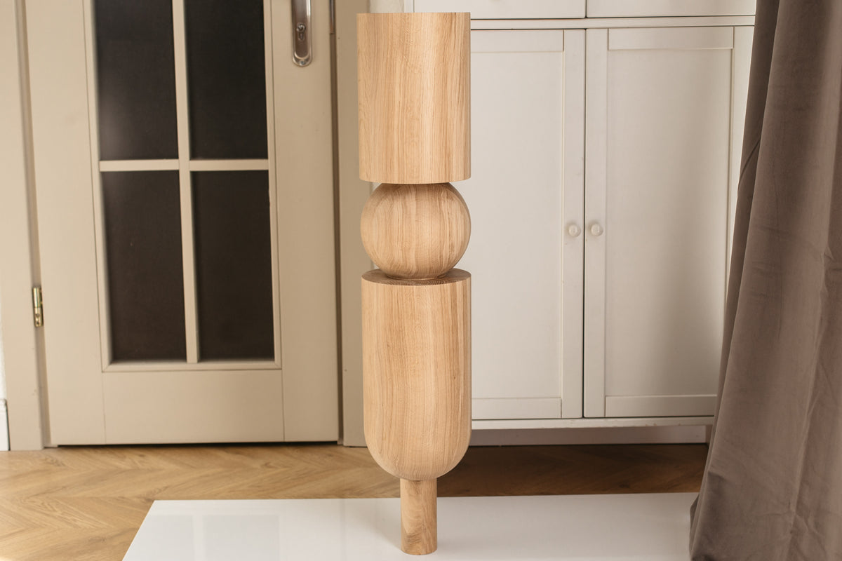 A tall, sculptural wooden floor lamp with a natural light oak finish, featuring stacked cylindrical and spherical segments, shown standing on a white surface in a home interior.