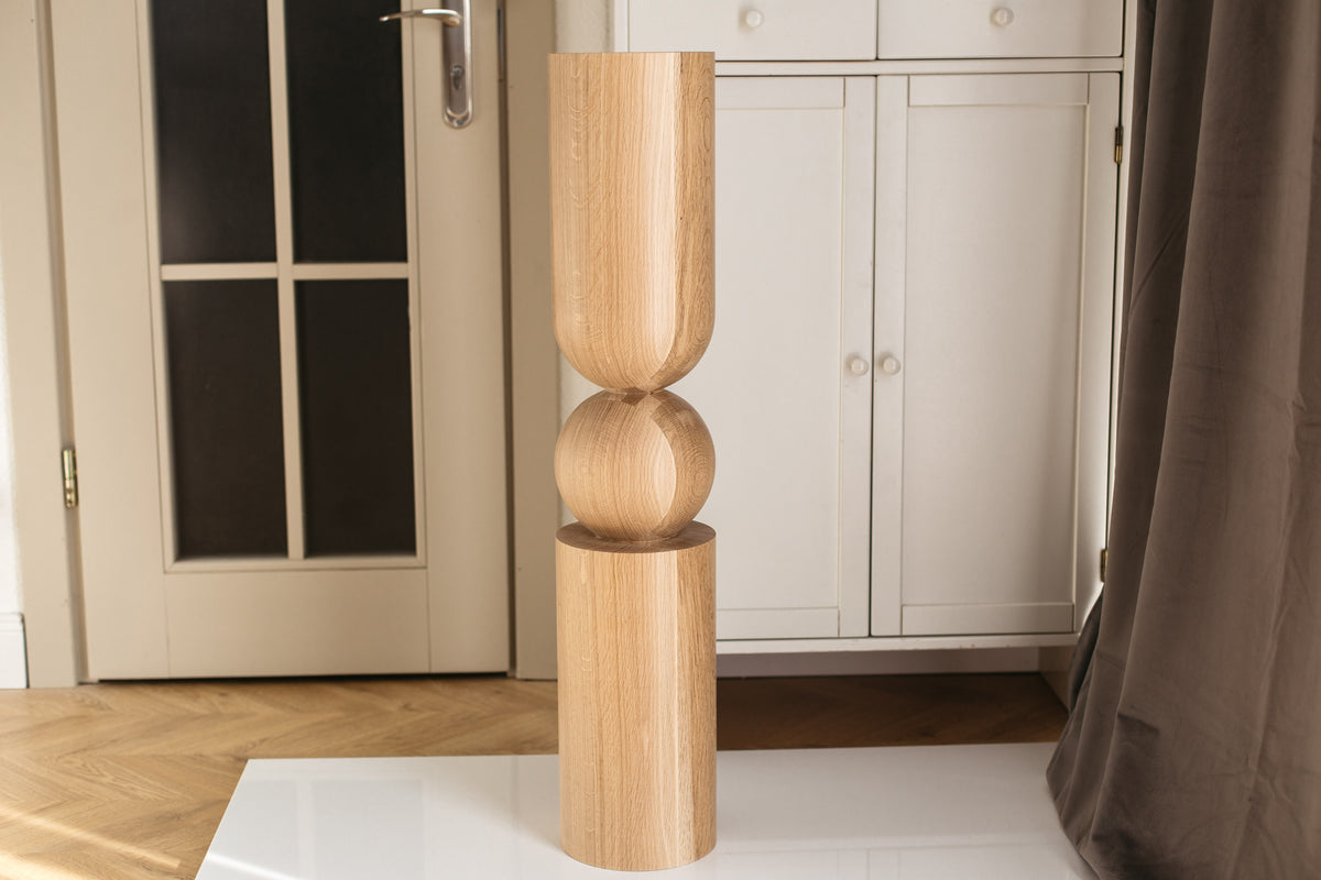 A tall, minimalist wooden sculptural lamp or decorative stand with a cylindrical top, a rounded middle section, and a flared base, shown indoors on a white table in front of white cabinets.
